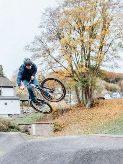 Een BMX-rijder springt over een ramp in een park. Op de achtergrond zijn bomen met herfstkleuren te zien.
