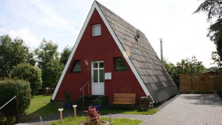 Een rood, driehoekig vakantiehuis met een schuin dak. Omgeven door een groene tuin en een terras.