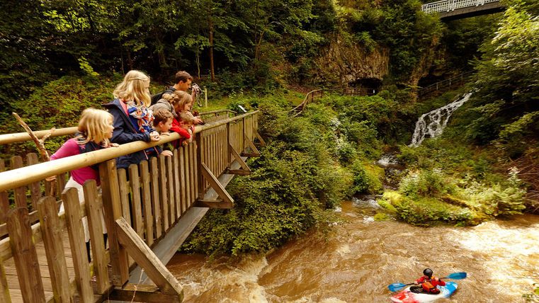 Een groep kinderen en volwassenen staat op een houten brug en kijkt naar een kajakker in het woeste water. Op de achtergrond zijn bomen en een kleine waterval te zien.