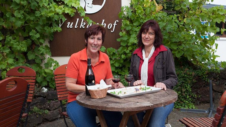 Zwei Frauen sitzen an einem Holztisch im Freien. Sie genießen Getränke und Essen vor einer Wand mit Weinreben und einem Schild.