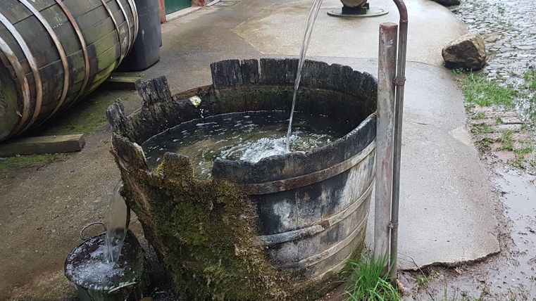 Ein alter Holzfass mit fließendem Wasser steht auf einer gepflasterten Fläche. Umgeben von Steinen und Gras vermittelt die Szene einen rustikalen Charme.