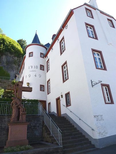 Weißes Gebäude mit Turm und roten Fensterrahmen, Treppe führt hinauf. Vor dem Gebäude steht eine Statue mit Kreuz.