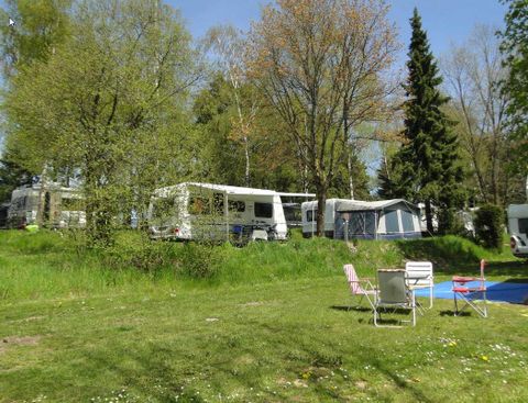 Ein Campingplatz mit Wohnmobilen und Zelten umgeben von grünen Bäumen. Im Vordergrund stehen zwei Gartenstühle auf einer grünen Wiese.