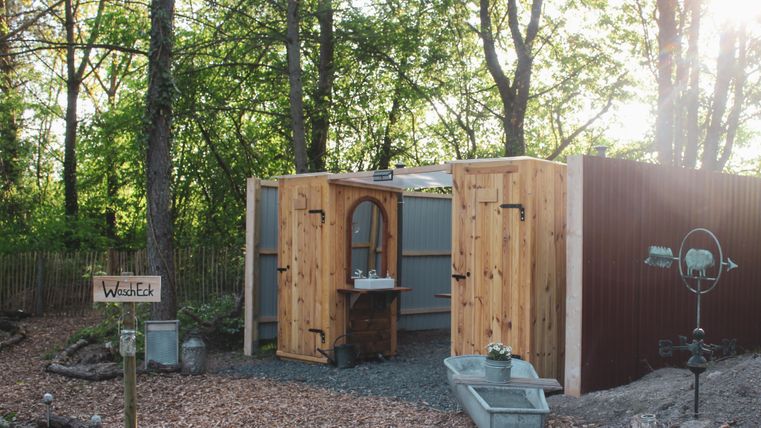 Een rustieke, vrijstaande badkamer in het bos met houten panelen en een spiegel. Er is een rustieke wastafelinstallatie en een pad van houtschors.