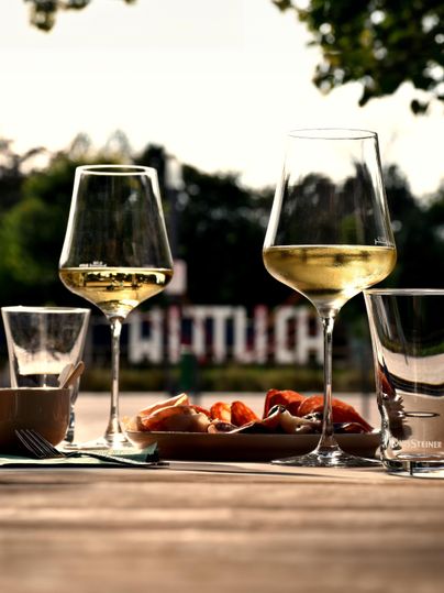 A table with several glasses of white wine and spirits as well as a plate of snacks. In the background, there are trees and a sign visible.