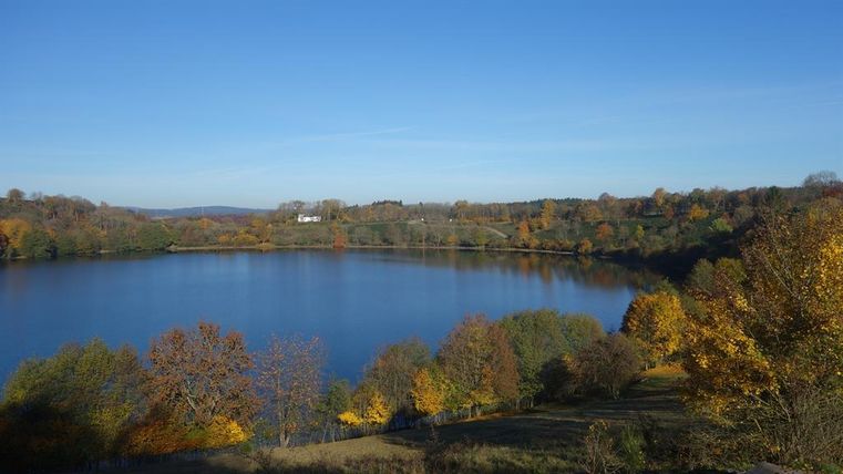 Ein ruhiger See umgeben von bunten Bäumen im Herbst. Der klare Himmel spiegelt sich im Wasser wider.