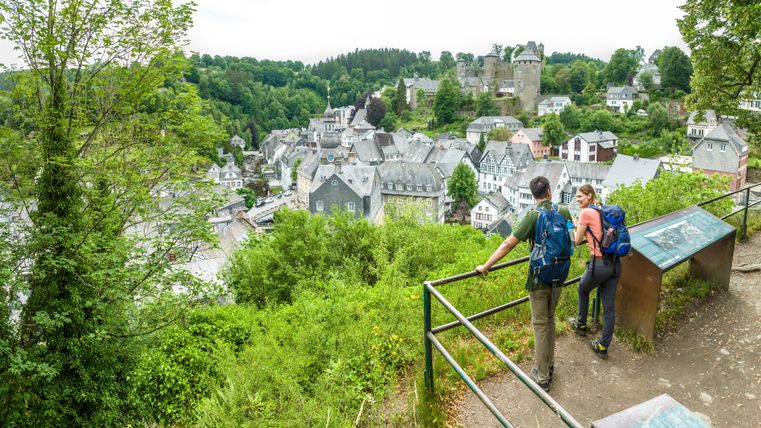 Eine malerische Aussicht auf ein kleines Dorf mit historischen Gebäuden und einer Burg im Hintergrund. Zwei Wanderer genießen die Aussicht von einem Aussichtspunkt umgeben von grüner Natur.