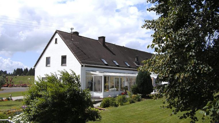 Ein helles Haus mit einem Garten und einer Terrasse. Der Himmel ist bewölkt und die Umgebung ist grün.