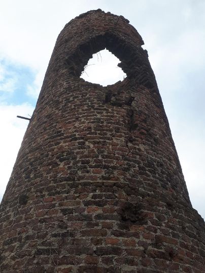 Een oude, vervallen toren van rode bakstenen met een groot gat aan de top. De lucht is bewolkt en het ziet er somber uit.