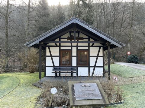 Ein kleines, traditionelles Fachwerkhaus in einer grünen Umgebung. Im Vordergrund steht eine Gedenktafel.