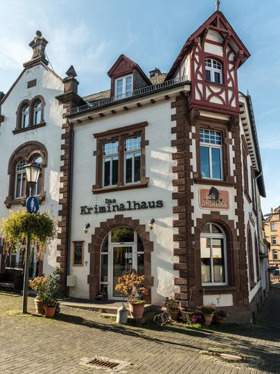 Historisches Kriminalhaus in Hillesheim mit dem Café Sherlock. Die Fassade ist weiß mit roten Akzenten. Vor dem Eingang stehen Pflanzen.