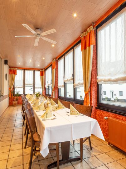 Een licht restaurant met een lange tafel die gedekt is voor een feest. Grote ramen zorgen voor veel daglicht en een vriendelijke sfeer.