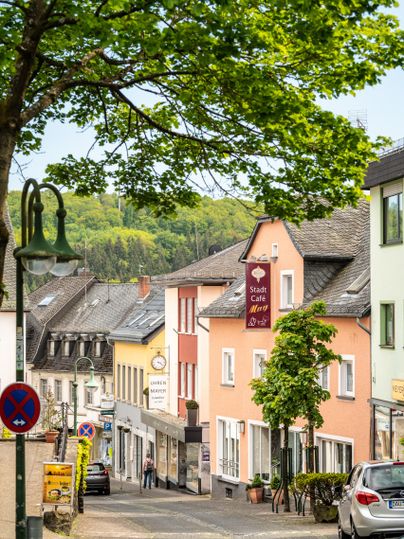 Eine charmante Straße mit bunten Häusern und einer klaren Sicht auf die Natur im Hintergrund. Ein Café und freundliche Bäume verleihen der Szene eine einladende Atmosphäre.