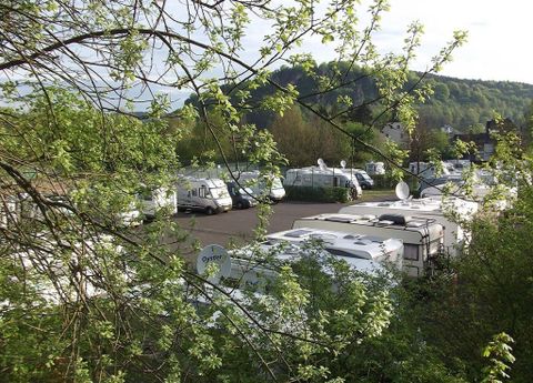 Ein Wohnmobilstellplatz mit mehreren geparkten Wohnmobilen, umgeben von grünen Bäumen und Hügeln im Hintergrund.