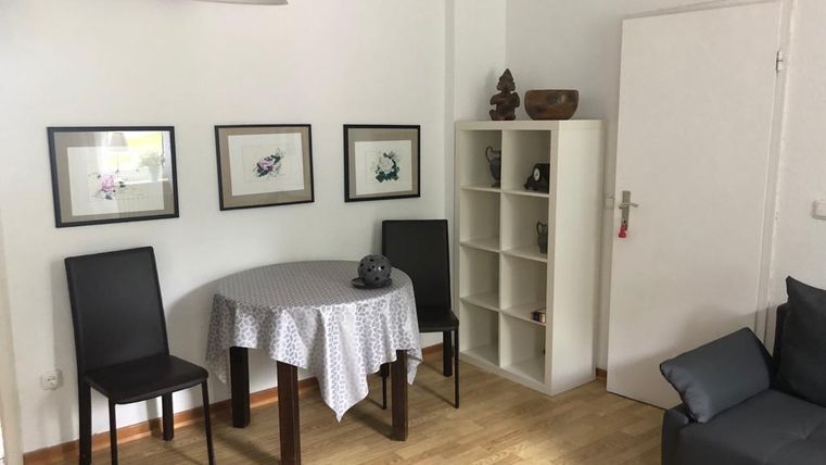 Ein gemütlicher Raum mit einem runden Tisch und zwei Stühlen. An der Wand hängen Bilder, und es gibt ein Regal in der Ecke.