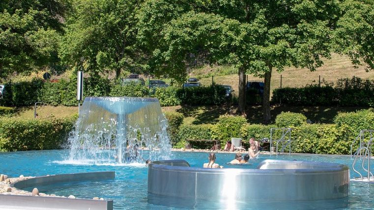 Ein entspannendes Schwimmbecken mit einem Wasserfall in einem grünen Umfeld. Menschen genießen die Sonne am Rand des Beckens.