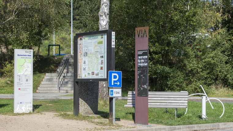 An information stand with maps and signposts in a park. In the foreground, there is a parking sign and a bench.