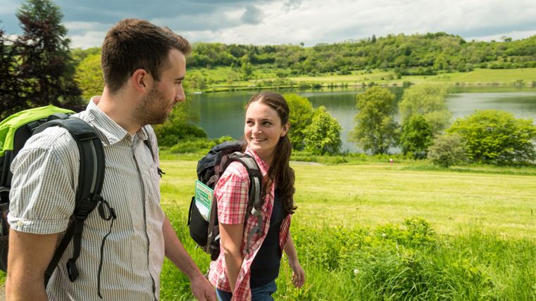 Two hikers at Schalkenmehrener Maar with rucksacks, the lake and green hills in the background.