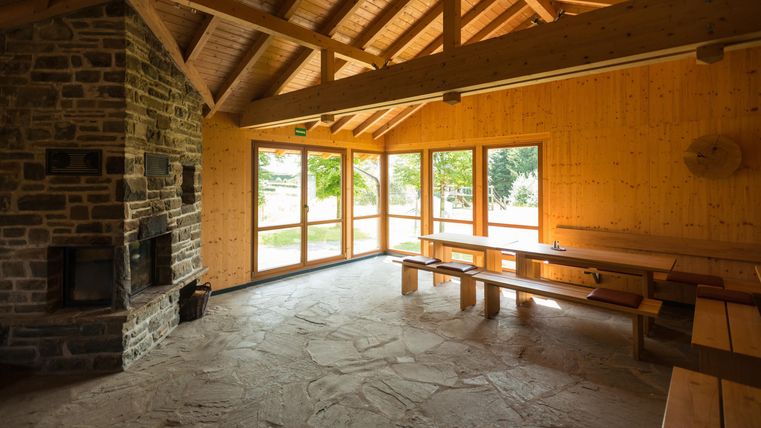 Ein gemütlicher Raum mit Holzwänden und großen Fenstern. In der Mitte stehen Holzbänke und ein Kamin aus Stein.