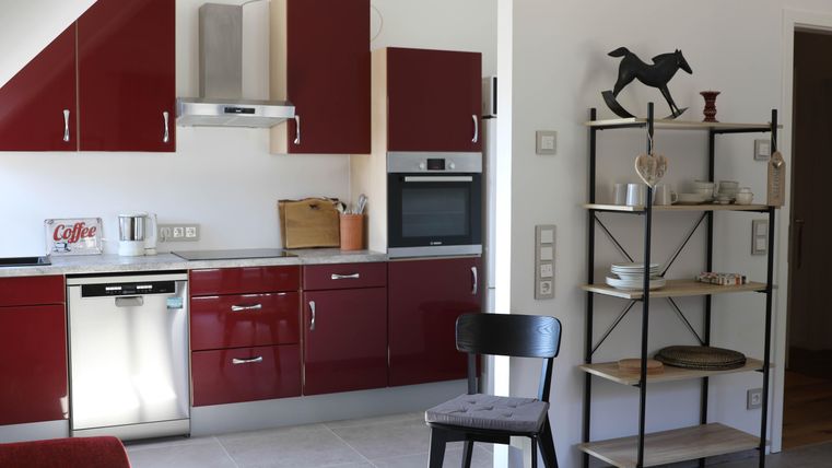 A modern kitchen with red cabinets and bright countertops. A shelf with dishes and decorations complements the space.