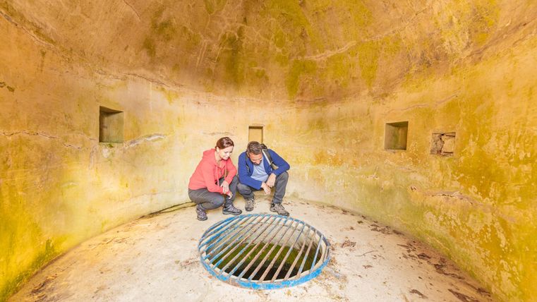 Zwei Personen betrachten einen Gitterrost in einem großen, leeren Raum mit einer gekrümmten Decke. Die Wände sind farblich verblasst und zeigen Moosansammlungen.