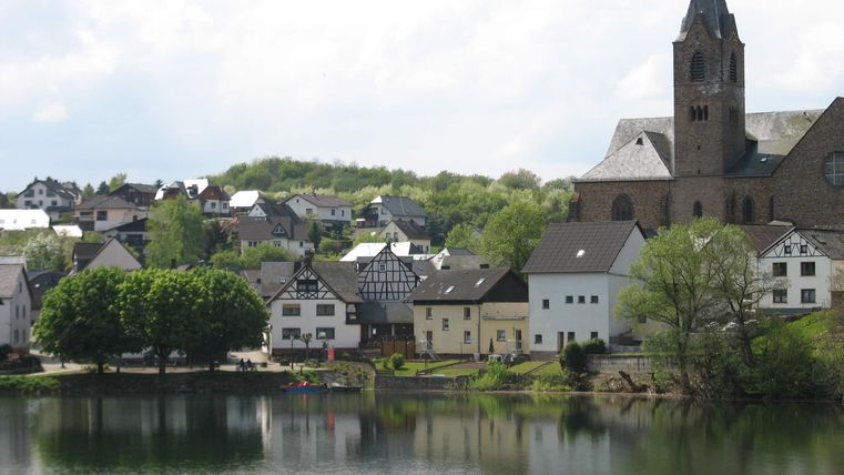 Eine malerische Stadt am Ufer eines Sees mit traditionellen Häusern und einer Kirche. Die Grünflächen und der klare Himmel sorgen für eine idyllische Atmosphäre.