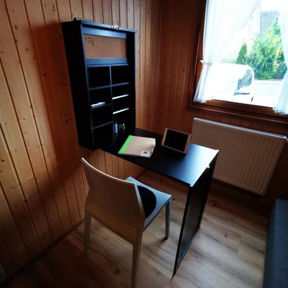 Een modern bureau met een witte stoel in een houten kamer. Op de tafel liggen schrijfgerei en een tablet, terwijl het licht door een raam naar binnen stroomt.