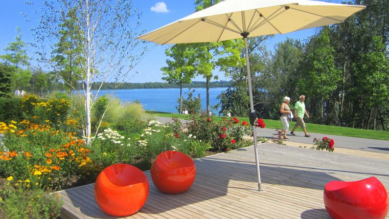 Een ontspannende buitenruimte met rode stoelen en een parasol. Op de achtergrond is het meer en een bloeiende tuinlandschap te zien.