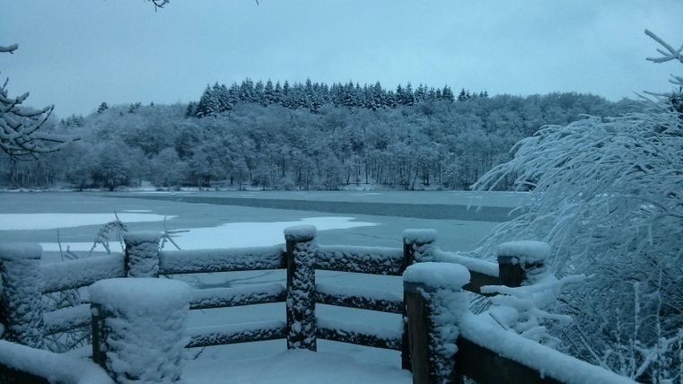 Eine verschneite Landschaft mit einem gefrorenen See und schneebedeckten Bäumen. Im Vordergrund ist eine Holzbalkonbrüstung zu sehen.