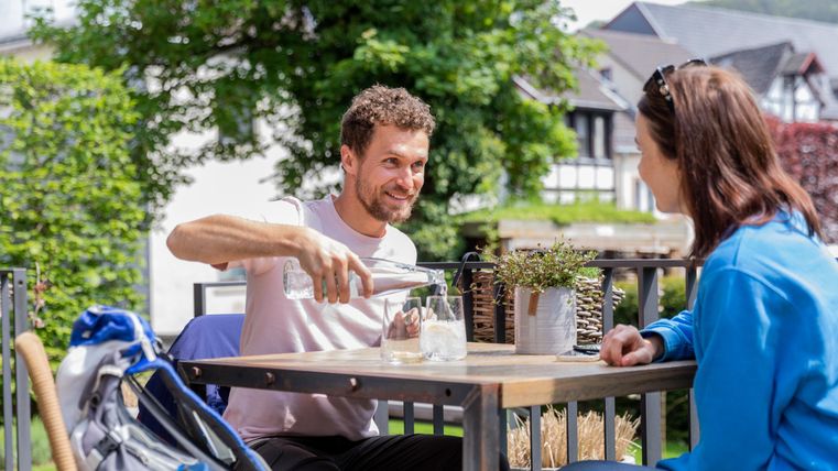 A man pours water into a glass while talking to a woman at an outdoor table. The surroundings are green and sunny, creating a relaxed atmosphere.