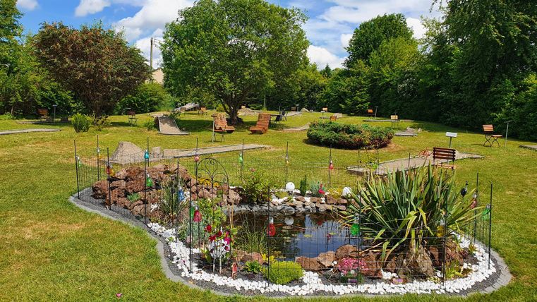 Ein schöner Garten mit einem Teich und bunten Blumen. Im Hintergrund sind Bäume und gemütliche Sitzgelegenheiten zu sehen.