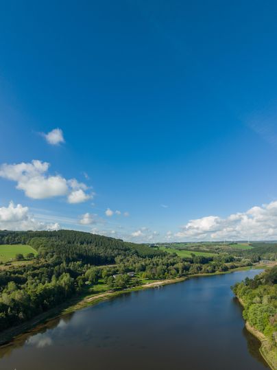 Man blickt von oben auf den Kronenburger See. Links und rechts der Wasserfläche sind sanft ansteigende Hügel die mit Laub und /oder Fichtenwälder überzogen sind. Zwischendurch liegen vereinzelt grüne Wiesen.