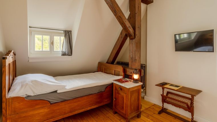 A cozy bedroom with a single bed and wooden beams. On the wall, there is a television and a wooden table.