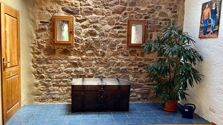 Ein einladender Raum mit einer Steinwand und zwei Bilderrahmen. In der Mitte steht eine dunkle Truhe und eine große Pflanzen steht in der Ecke.