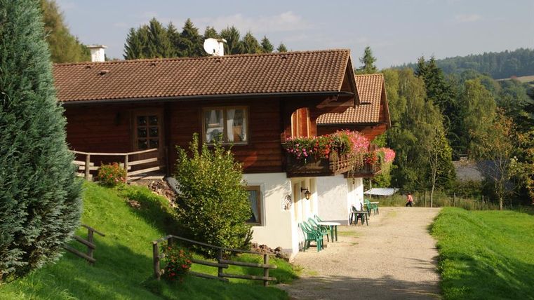 Ein gemütliches Holzhaus mit Balkon und Blumen in einer malerischen Landschaft. Der Weg führt sanft zu dem Haus, umgeben von Bäumen und Wiesen.
