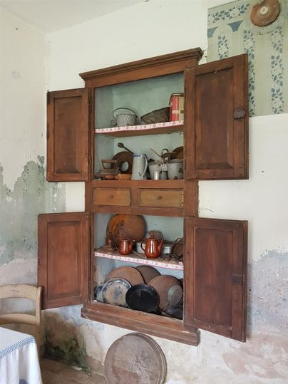 Ein antiker Schrank mit offenem Inhalt zeigt verschiedene Kochutensilien und Töpfe. Die Wände sind teilweise verputzt und verleihen dem Raum einen rustikalen Charme.