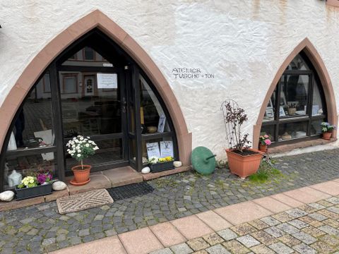 Ein einladendes Geschäft mit großen Fenstern in gotischem Stil. Draußen stehen blühende Pflanzen in Töpfen und das Ambiente ist freundlich und kreativ.