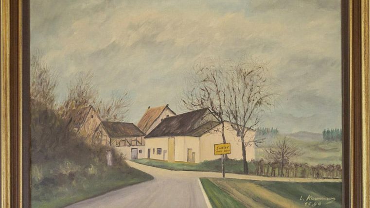 Eine ländliche Szene mit einem ruhigen Weg, der an einigen Häusern vorbeiführt. Im Hintergrund sind sanfte Hügel und ein grauer Himmel zu sehen.