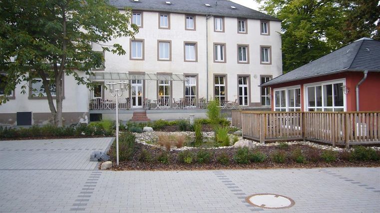 Ein Gebäude mit mehreren Fenstern, umgeben von einem gepflegten Garten. Es gibt einen Bereich mit Holzgeländer und Pflanzen.