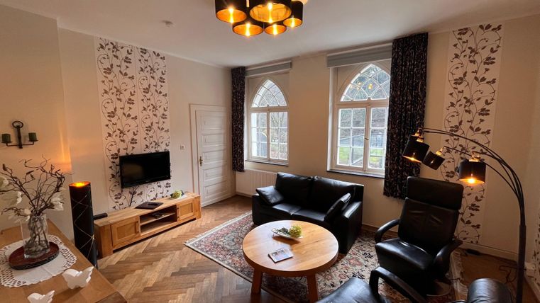 Ein gemütliches Wohnzimmer mit eleganter Einrichtung und großen Fenstern. Es gibt einen runden Tisch, bequeme Sessel und eine moderne Beleuchtung.