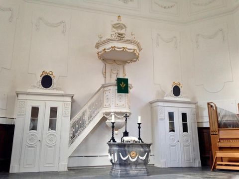 Ein eleganter Innenraum mit einer weiße Kanzel und zwei Deuren. Auf der linken Seite steht eine Orgel und die Wände sind schlicht verziert.