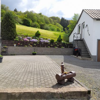 A quiet courtyard with a paved floor and a bright, inviting atmosphere. In the background, well-maintained flowers and green spaces can be seen.