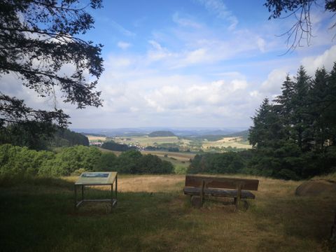 A beautiful view over the valley with gentle hills and wooded areas. In the foreground are a bench and an information table.