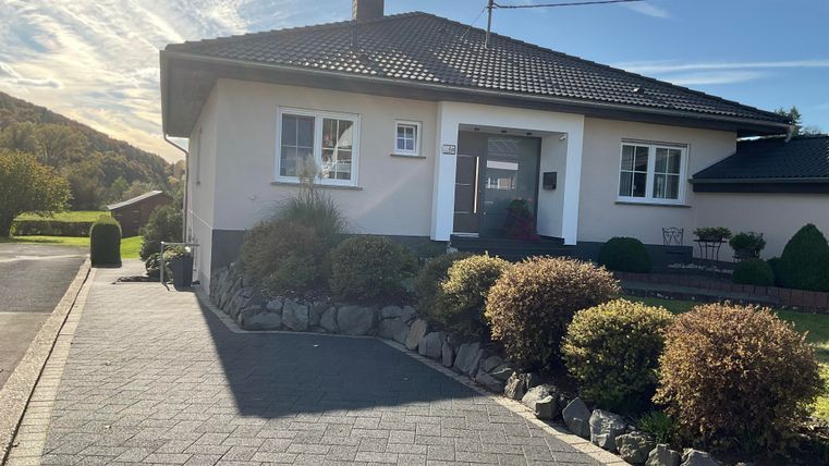 A well-maintained single-family home with a beautiful garden and a paved driveway. The surroundings are bordered by green hills.