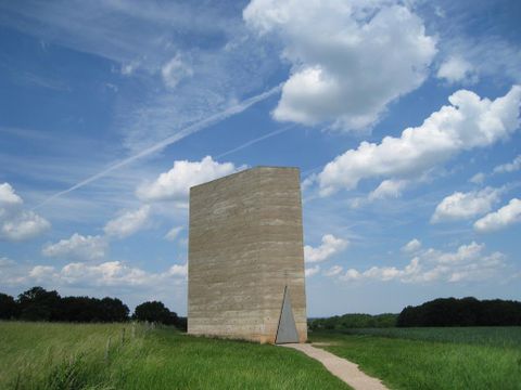 Ein großes, rechteckiges Bauwerk aus Beton steht auf einem grünen Feld unter einem klaren Himmel mit wenigen Wolken. Ein schmaler Weg führt zur Eingangstür.