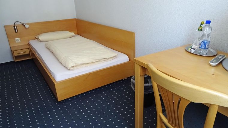 Ein einfaches Zimmer mit einem Einzelbett und einem Tisch. Auf dem Tisch steht eine Flasche Wasser und ein Telefon.