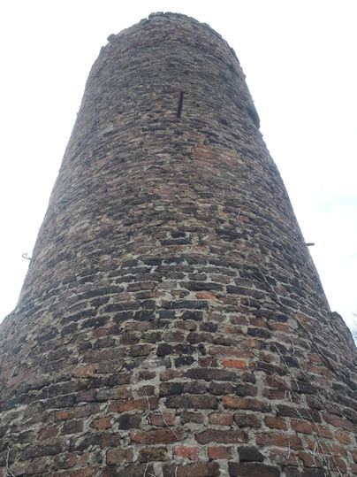 Een historisch stenen gebouw dat hoog de lucht in steekt. De toren heeft een rustieke structuur van baksteen en wordt omgeven door een grijs wolkenlandschap.