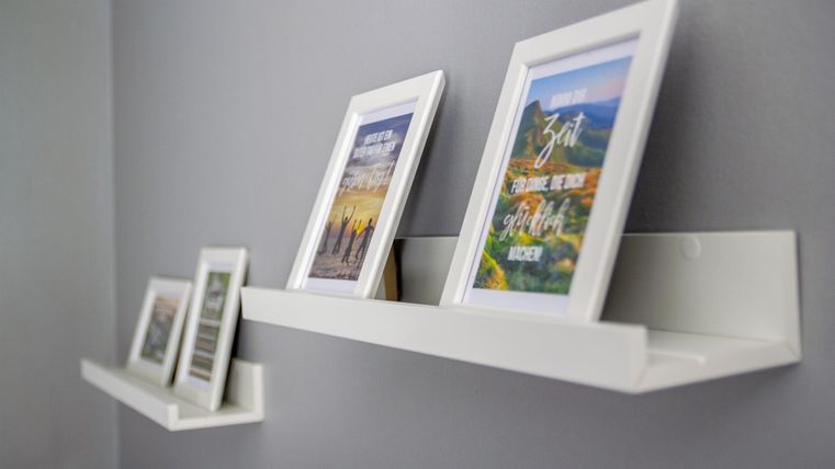Frames with pictures are on simple, white shelves against a gray wall. The pictures depict various motifs and give the room a personal touch.