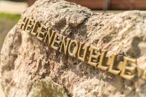 Ein Stein mit dem Schriftzug 'Helenenquelle' in goldenen Buchstaben, sonnig beleuchtet.
