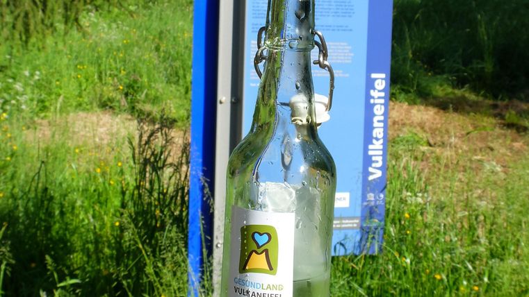 A glass bottle with a label is in the foreground, while an information sign is visible in the background. The surroundings consist of green grass and trees.
